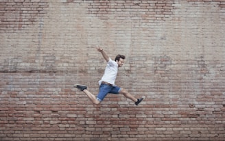 person in white shirt and blue denim jeans jumping beside brown walll
