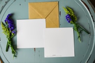 A brown kraft envelope and two blank, square-shaped white cards are laid out on a round, weathered, teal wooden surface. Sprigs of purple and green flowers are placed on either side of the cards and envelope.