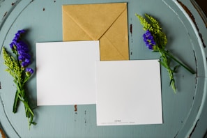 A brown kraft envelope and two blank, square-shaped white cards are laid out on a round, weathered, teal wooden surface. Sprigs of purple and green flowers are placed on either side of the cards and envelope.
