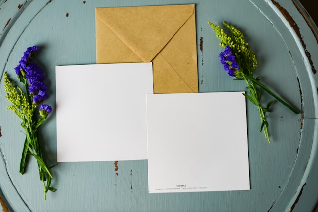 A brown kraft envelope and two blank, square-shaped white cards are laid out on a round, weathered, teal wooden surface. Sprigs of purple and green flowers are placed on either side of the cards and envelope.