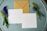 A brown kraft envelope and two blank, square-shaped white cards are laid out on a round, weathered, teal wooden surface. Sprigs of purple and green flowers are placed on either side of the cards and envelope.