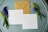 A brown kraft envelope and two blank, square-shaped white cards are laid out on a round, weathered, teal wooden surface. Sprigs of purple and green flowers are placed on either side of the cards and envelope.