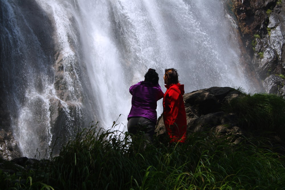 two persons standing on grass looking at the waterfalls
