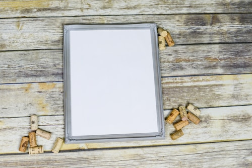 A blank framed canvas is surrounded by scattered wine corks on a rustic wooden surface. The scene has a vintage and artistic vibe, with corks placed randomly around the frame, suggesting a casual, creative atmosphere.