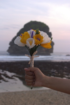 A hand is holding a bouquet of artificial flowers with bright yellow, purple, and white petals, wrapped with a light-colored ribbon. In the background, there is a large rock formation on the shore with a gentle ocean wave rolling in and a cloudy sky, suggesting a coastal setting during dawn or dusk.