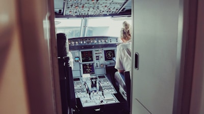Inside the cockpit of an aircraft, two pilots are seated. The cockpit is filled with numerous controls, screens, and displays on the dashboard. There is ambient lighting, and the pilots are focused on the flight controls. The area is compact and filled with technical equipment.