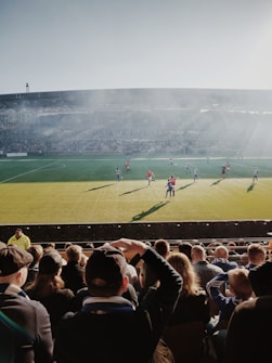 A crowd of spectators watches a football match in a stadium under a clear sky. Players in red and blue jerseys are actively engaged in the game on a green field, while sunlight creates long shadows. The stadium is filled with cheering fans, with some standing and others seated, capturing the vibrant atmosphere.