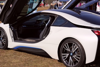 A sleek white sports car with gull-wing doors open, revealing the interior. The car is parked on grass, with a display in the background showcasing balloons and various items. The car's modern design features aerodynamic lines and futuristic details.