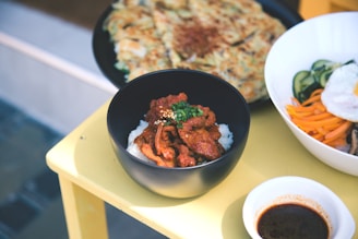 A close-up of a meal on a yellow table featuring a black bowl containing rice with spicy red meat garnished with scallions and sesame seeds. In the background, there is a partially visible plate of pancake-like food, and a white bowl containing cucumber slices, carrots, and a fried egg. A small bowl of dark sauce sits next to the main dish.