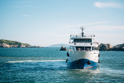 A ferry boat is moving across the ocean with clear blue skies above. Rocky islands can be seen in the background.
