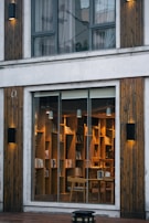 A cozy and inviting interior library with wooden bookshelves filled with books, visible through a large glass window. Warm lighting creates an intimate atmosphere, with wooden chairs arranged around small tables. The exterior features vertical wooden paneling and modern light fixtures.