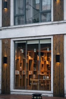 A cozy and inviting interior library with wooden bookshelves filled with books, visible through a large glass window. Warm lighting creates an intimate atmosphere, with wooden chairs arranged around small tables. The exterior features vertical wooden paneling and modern light fixtures.