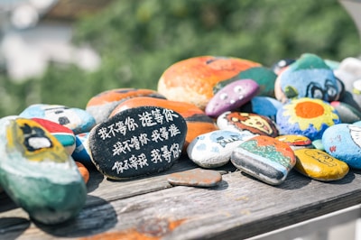 A collection of smooth, brightly painted rocks is arranged on a wooden surface. Each rock features unique designs such as flowers, abstract patterns, animals, and faces. One prominent rock displays writing in Chinese characters, adding a personal or cultural element to the scene. The colorful stones are illuminated by soft sunlight, creating a cheerful and artistic atmosphere.