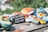 A collection of smooth, brightly painted rocks is arranged on a wooden surface. Each rock features unique designs such as flowers, abstract patterns, animals, and faces. One prominent rock displays writing in Chinese characters, adding a personal or cultural element to the scene. The colorful stones are illuminated by soft sunlight, creating a cheerful and artistic atmosphere.