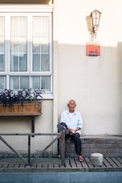 An elderly man is sitting on a wooden bench outside a building with large windows and a flower box. He is wearing a light blue shirt and dark pants. A lamp is mounted on the wall above him, and there is a red plaque on the wall. The scene is lit by natural sunlight.