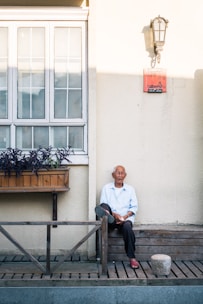 An elderly man is sitting on a wooden bench outside a building with large windows and a flower box. He is wearing a light blue shirt and dark pants. A lamp is mounted on the wall above him, and there is a red plaque on the wall. The scene is lit by natural sunlight.