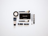 A collection of makeup products arranged neatly on a white surface. Items include multiple brushes, eyeliner, foundation, a makeup palette, and lipstick. The products are evenly spaced and organized, presenting a clean and minimalist aesthetic.