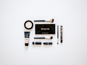 A collection of makeup products arranged neatly on a white surface. Items include multiple brushes, eyeliner, foundation, a makeup palette, and lipstick. The products are evenly spaced and organized, presenting a clean and minimalist aesthetic.