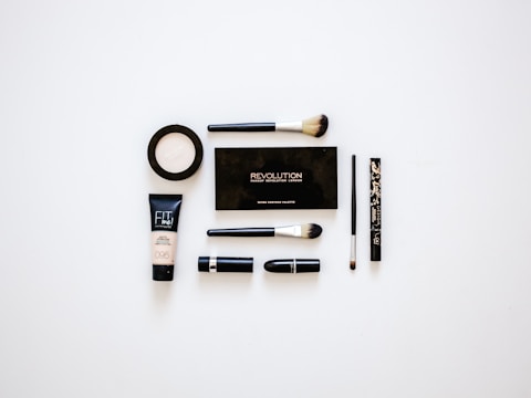 A collection of makeup products arranged neatly on a white surface. Items include multiple brushes, eyeliner, foundation, a makeup palette, and lipstick. The products are evenly spaced and organized, presenting a clean and minimalist aesthetic.