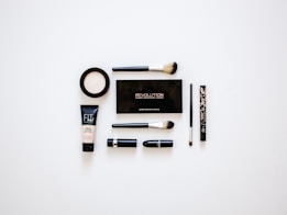 A collection of makeup products arranged neatly on a white surface. Items include multiple brushes, eyeliner, foundation, a makeup palette, and lipstick. The products are evenly spaced and organized, presenting a clean and minimalist aesthetic.