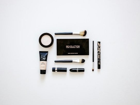 A collection of makeup products arranged neatly on a white surface. Items include multiple brushes, eyeliner, foundation, a makeup palette, and lipstick. The products are evenly spaced and organized, presenting a clean and minimalist aesthetic.