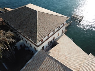 Aerial view of a large, tiled-roof building adjacent to a body of water. The structure has a symmetrical, triangular roof design, with numerous windows lined along its white walls. Adjacent to the building’s edge is a small pier or dock extending into the calm, reflective water. Palm trees are visible in one corner near the building.