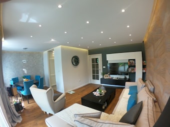 The interior of a modern living room with a sectional sofa, a beige armchair, and a large black coffee table. A flat-screen TV is mounted on a wall with white and dark wood shelves filled with books and decorations. On the left side, there's a dining area with a round black table surrounded by blue upholstered chairs, with a centerpiece of fruit. The walls have a textured design, and recessed lighting is embedded in the ceiling. The floor is wooden, and the space is well lit with natural and artificial lighting.