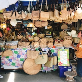 A market stall displays a variety of woven baskets and handbags in different shapes and sizes. The baskets are made from natural materials and are predominantly beige in color. Two people are visible, one in the background and another on the right side, who appears to be interacting with the display. The stall is densely packed, creating a rustic and handmade vibe.