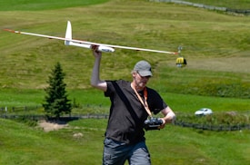 A man is holding a large model glider with one hand while operating a remote control in the other hand. He is wearing a cap, sunglasses, and a casual outfit. The background features a lush green landscape with a single evergreen tree, a small car, and a cable car in the distance.