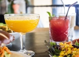 A close-up of a table set with colorful drinks and a fresh salad. A yellow-orange drink is served in an elegantly patterned glass, next to a red drink with a straw and mint garnish. The foreground features a vibrant salad with corn, greens, and other vegetables.
