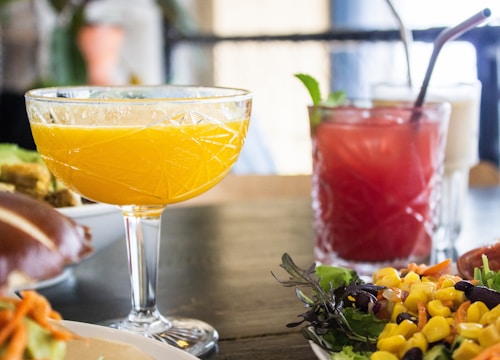 A close-up of a table set with colorful drinks and a fresh salad. A yellow-orange drink is served in an elegantly patterned glass, next to a red drink with a straw and mint garnish. The foreground features a vibrant salad with corn, greens, and other vegetables.