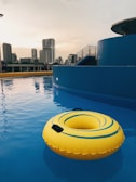 A rooftop swimming pool with a large yellow inflatable ring floating on the calm water. The background features a city skyline with several tall buildings under a cloudy sky. The deck area is equipped with modern stairs and a railing.