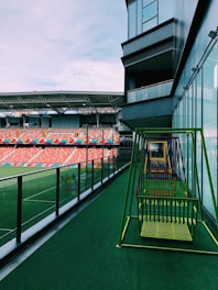 A modern sports stadium with tiered seating and colorful rows, viewed from a balcony area featuring a series of colorful swings. The structure includes a glass facade along the side of the balcony, reflecting a part of the sky and stadium. The seating area is mostly empty, with a vast stretch of green artificial turf visible on the field.