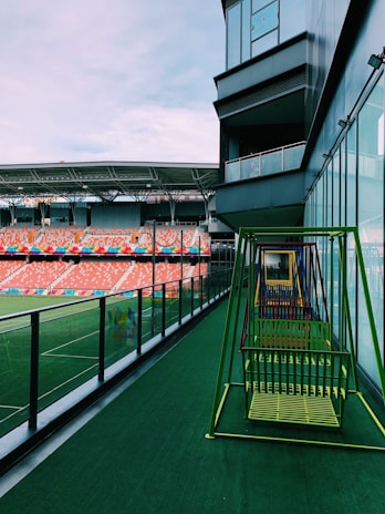 A modern sports stadium with tiered seating and colorful rows, viewed from a balcony area featuring a series of colorful swings. The structure includes a glass facade along the side of the balcony, reflecting a part of the sky and stadium. The seating area is mostly empty, with a vast stretch of green artificial turf visible on the field.