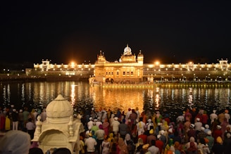 A group of people gathers near a large, illuminated golden temple reflected in the water, creating a shimmering effect at night. The temple is surrounded by a larger white building complex with lights that enhance its majestic appearance. A calm, sacred atmosphere is conveyed by the gathering.