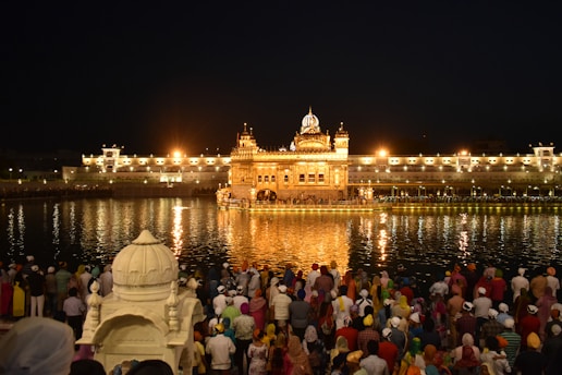 A group of people gathers near a large, illuminated golden temple reflected in the water, creating a shimmering effect at night. The temple is surrounded by a larger white building complex with lights that enhance its majestic appearance. A calm, sacred atmosphere is conveyed by the gathering.