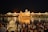 A group of people gathers near a large, illuminated golden temple reflected in the water, creating a shimmering effect at night. The temple is surrounded by a larger white building complex with lights that enhance its majestic appearance. A calm, sacred atmosphere is conveyed by the gathering.