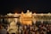 A group of people gathers near a large, illuminated golden temple reflected in the water, creating a shimmering effect at night. The temple is surrounded by a larger white building complex with lights that enhance its majestic appearance. A calm, sacred atmosphere is conveyed by the gathering.