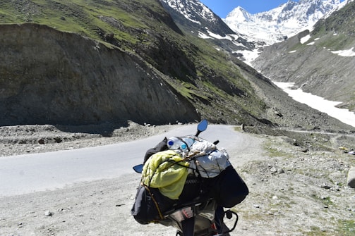 A scenic mountain landscape featuring a winding road that passes through rocky terrain with patches of greenery. In the foreground, a motorcycle is loaded with travel gear, including a yellow bag and a rolled-up sleeping bag. Snow-capped peaks loom in the distance under a clear blue sky.