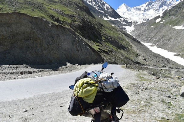 A scenic mountain landscape featuring a winding road that passes through rocky terrain with patches of greenery. In the foreground, a motorcycle is loaded with travel gear, including a yellow bag and a rolled-up sleeping bag. Snow-capped peaks loom in the distance under a clear blue sky.