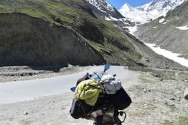 A scenic mountain landscape featuring a winding road that passes through rocky terrain with patches of greenery. In the foreground, a motorcycle is loaded with travel gear, including a yellow bag and a rolled-up sleeping bag. Snow-capped peaks loom in the distance under a clear blue sky.
