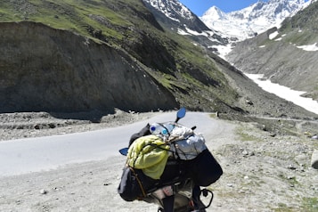 A scenic mountain landscape featuring a winding road that passes through rocky terrain with patches of greenery. In the foreground, a motorcycle is loaded with travel gear, including a yellow bag and a rolled-up sleeping bag. Snow-capped peaks loom in the distance under a clear blue sky.