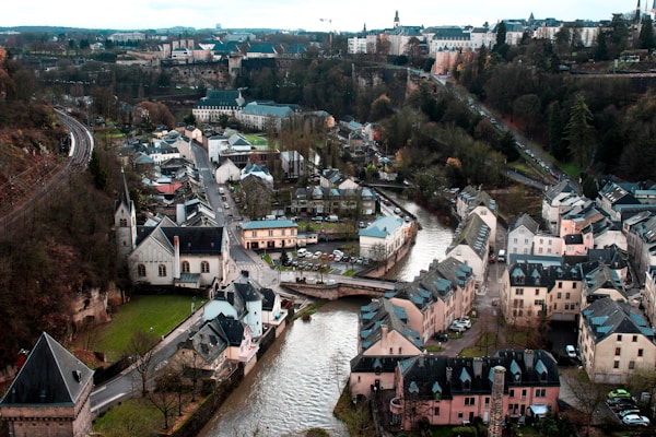A picturesque urban landscape featuring a historic European town with charming, pastel-colored buildings nestled along a winding river. Narrow streets and a small bridge over the river enhance the town's quaint atmosphere. The surrounding area is lush with trees and greenery, and railway tracks can be seen to the left. The town is encircled by distant buildings and a hilly terrain in the background.