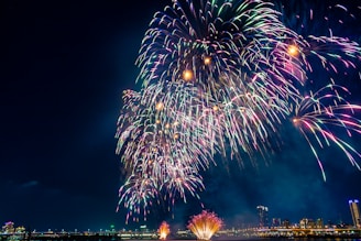 Vibrant fireworks burst in various colors against a clear night sky, illuminating a cityscape in the background. The fireworks display features large, colorful bursts that spread across the sky, creating a festive and celebratory atmosphere.