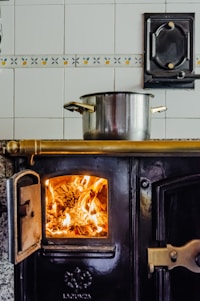 A vintage wood-burning stove with a bright fire glowing inside the open compartment. A shiny metal pot is placed on top, resting on a brass rod. The background features ceramic tiles with a decorative pattern, and the stove has ornate handles and details.