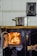 A vintage wood-burning stove with a bright fire glowing inside the open compartment. A shiny metal pot is placed on top, resting on a brass rod. The background features ceramic tiles with a decorative pattern, and the stove has ornate handles and details.