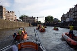Several orange rescue boats are navigating a canal bordered by historic buildings and a stone wall. People on the boats are wearing yellow vests and appear to be engaged in an organized activity. The scene includes a metal bridge and a distant view of more city structures.