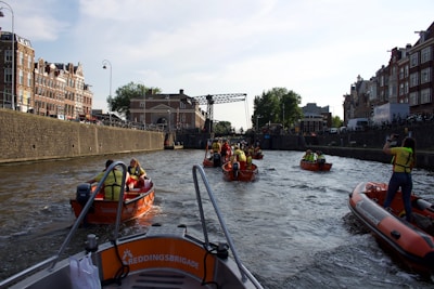Several orange rescue boats are navigating a canal bordered by historic buildings and a stone wall. People on the boats are wearing yellow vests and appear to be engaged in an organized activity. The scene includes a metal bridge and a distant view of more city structures.