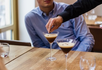 A person in a dark clothing sleeve is gently placing an item into a martini glass filled with a coffee-colored beverage, likely espresso martini, on a wooden table. Another person in a light blue shirt is seated nearby in a relaxed setting, with another identical drink on the table. The scene suggests a casual or social gathering.