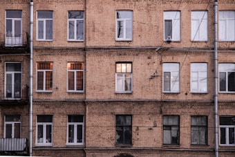 A multi-story apartment building with a brick facade features multiple windows, some of which are illuminated, suggesting habitation. The structure has a worn and aged appearance, with visible pipes and electrical wires running across its surface.